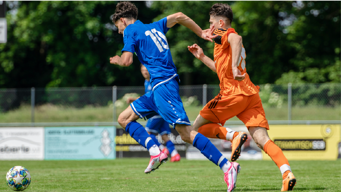 Two male soccer players in action on the field, one in blue and the other in orange, competing for the ball during a sunny day.